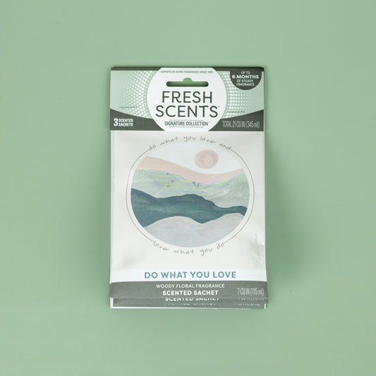The Fresh Scents Do What You Love - Sachet 3pk features a circular landscape of mountains, sun, and sky. This long-lasting flameless room freshener is shown on a plain light green background.