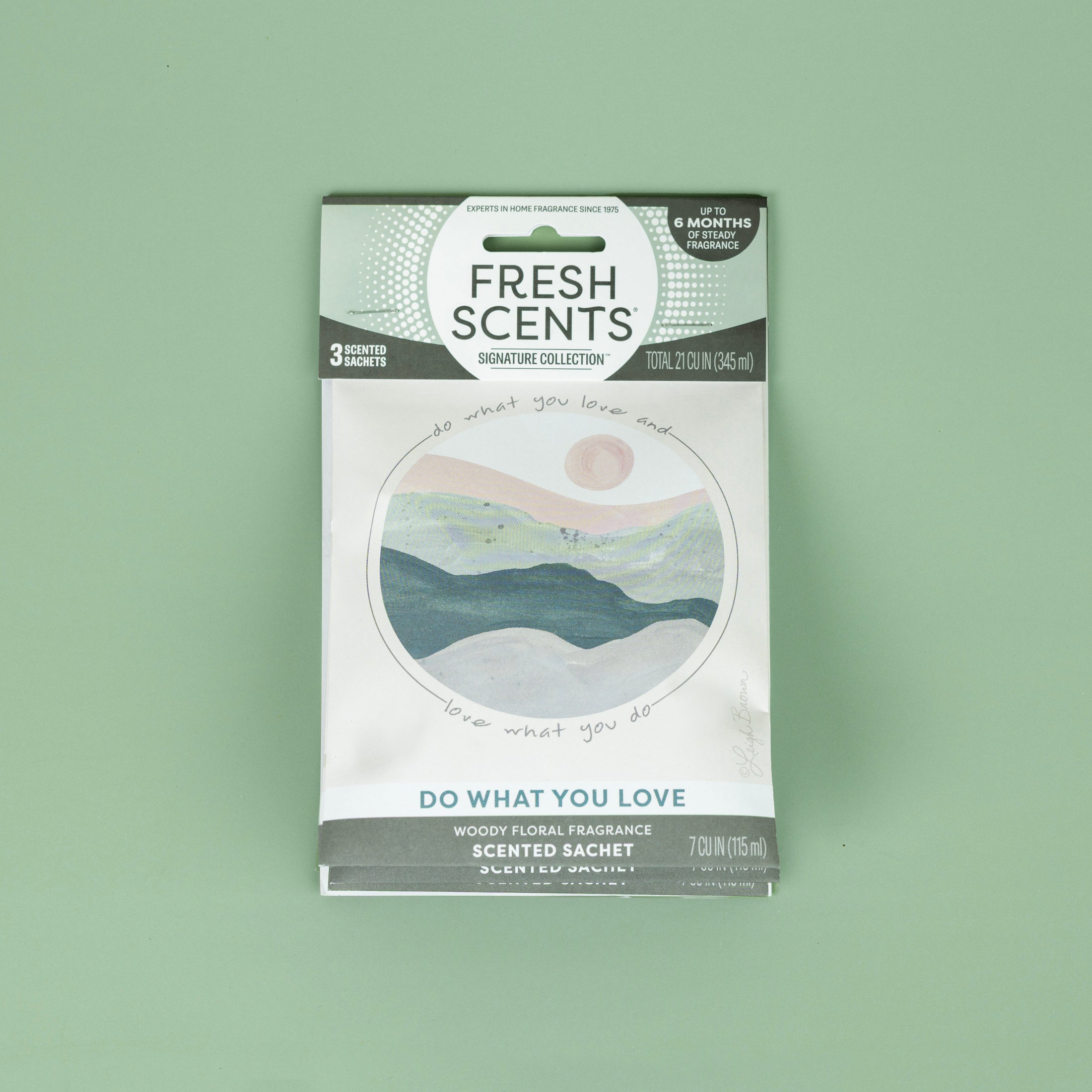 The Fresh Scents Do What You Love - Sachet 3pk features a circular landscape of mountains, sun, and sky. This long-lasting flameless room freshener is shown on a plain light green background.
