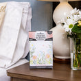 Load image into Gallery viewer, A box of Fresh Scents Baby Powder - Sachet 3pk sits on a wooden nightstand by a white lamp and daisies, providing a long-lasting air freshener beside a bed with crisp white sheets and pillows.
