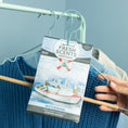Load image into Gallery viewer, A hand hangs a Fresh Scents Watermark - Sachet 3pk, a long-lasting scented sachet, on a clothes hanger with blue and beige clothing against a light blue background.