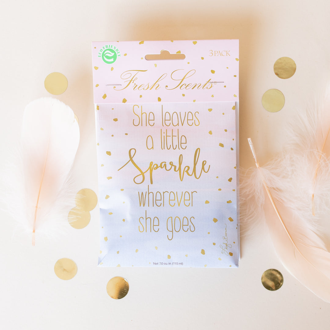A pastel sachet with gold dots and text reading She leaves a little sparkle wherever she goes, surrounded by white feathers and gold confetti on a light background.