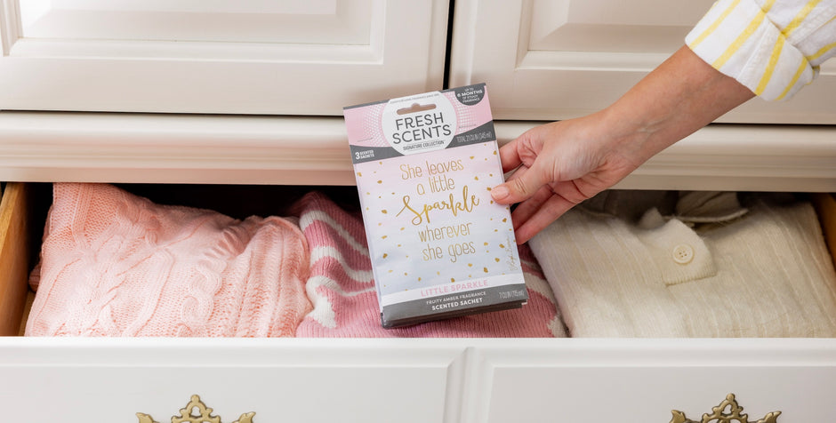 A hand places a Fresh Scents sachet labeled She leaves a little sparkle wherever she goes into a dresser drawer with neatly folded pink and white sweaters.