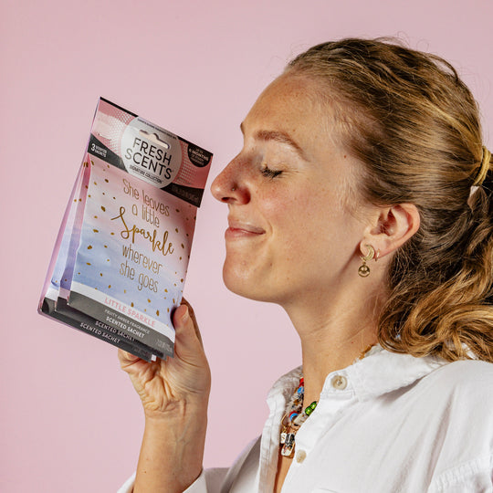 A woman with light hair smiles with her eyes closed, holding a Fresh Scents packet up to her nose. The packet reads, She leaves a little sparkle wherever she goes. The background is a solid light pink.