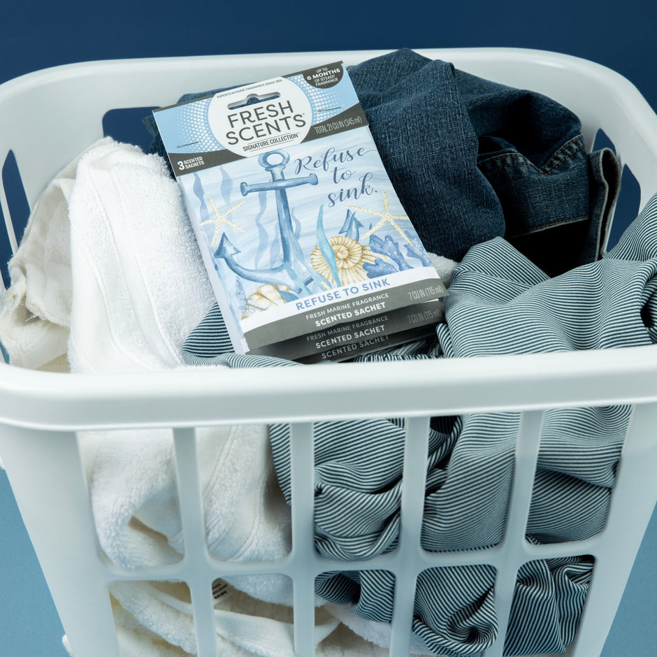 A white laundry basket filled with folded blue jeans, a striped shirt, and white towels, with a “Fresh Scents” scented sachet labeled “Refuse to Sink” placed on top. The background is blue.
