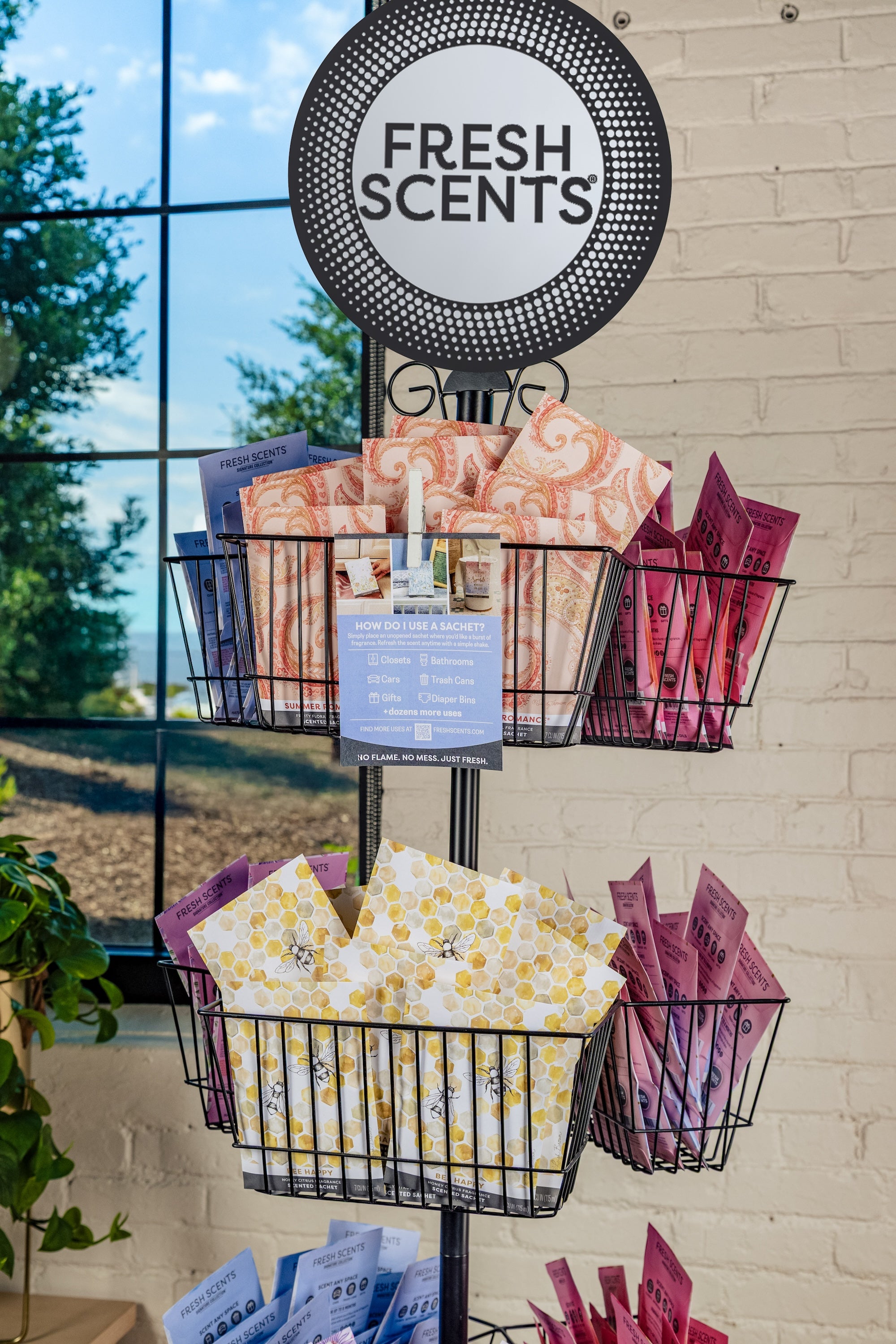 A black wire display rack holds colorful packets labeled Fresh Scents, arranged in three rows, with a round sign on top and a window showing trees and brick wall in the background.