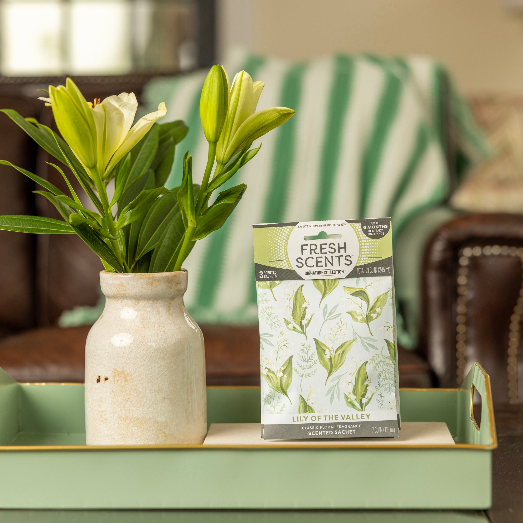 A ceramic vase with white lilies and a Fresh Scents Lily of the Valley - Sachet 3pk rests on a mint-green tray. In the background, a green and white striped blanket is draped over a brown couch.
