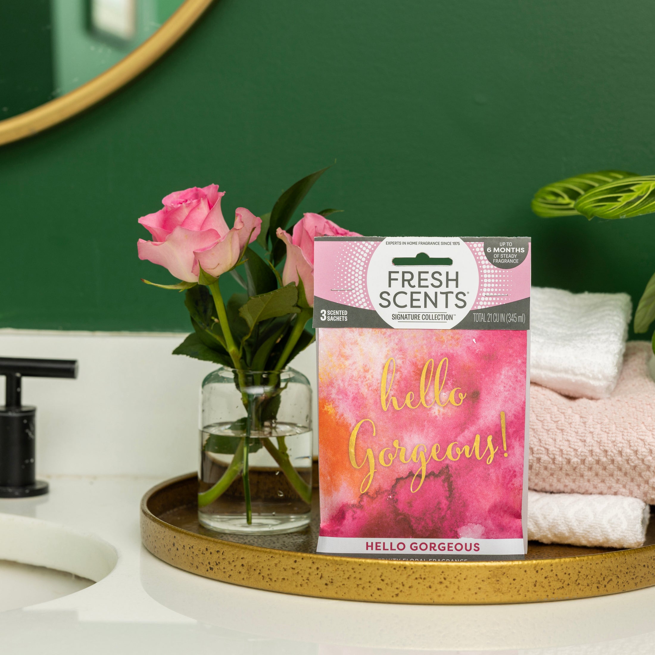 A bathroom counter with a gold tray displays a glass vase of pink roses, folded towels, and a Fresh Scents Hello Gorgeous - Sachet 3pk for long-lasting fragrance. In the background are a sink, green wall, and round mirror.