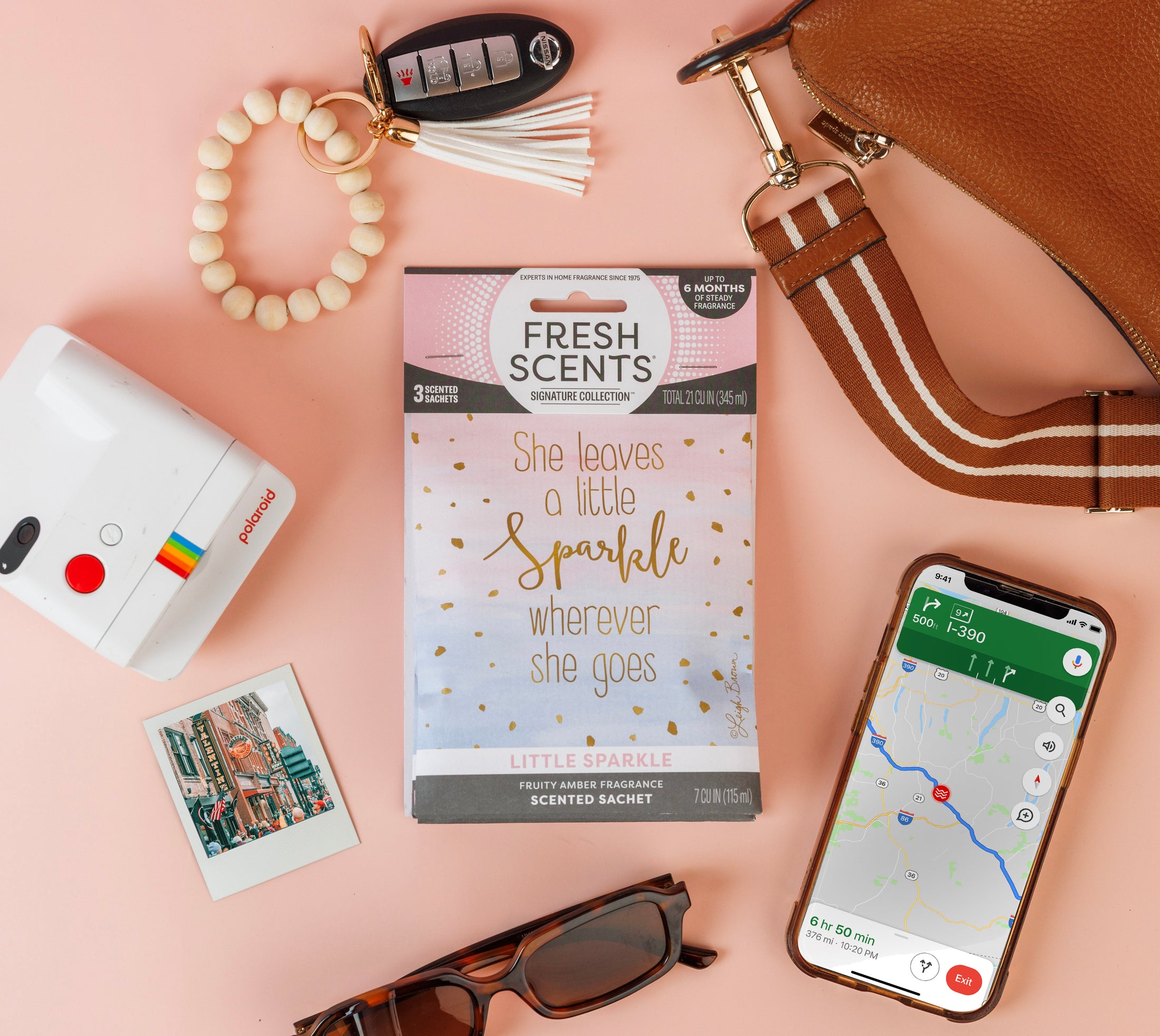 Flat lay featuring a purse, car keys, sunglasses, instant camera, photos, a Fresh Scents Little Sparkle - Sachet 3pk with “She leaves a little sparkle wherever she goes,” and a phone showing maps—all on a tan background.