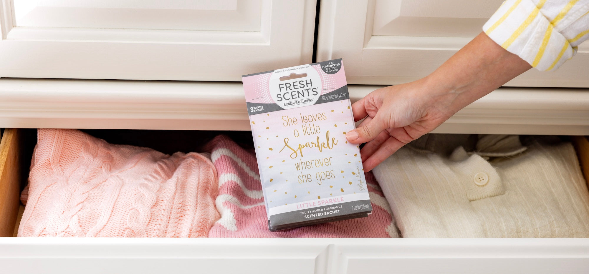 A hand places a Fresh Scents sachet labeled She leaves a little sparkle wherever she goes into a drawer filled with folded pink and cream sweaters.