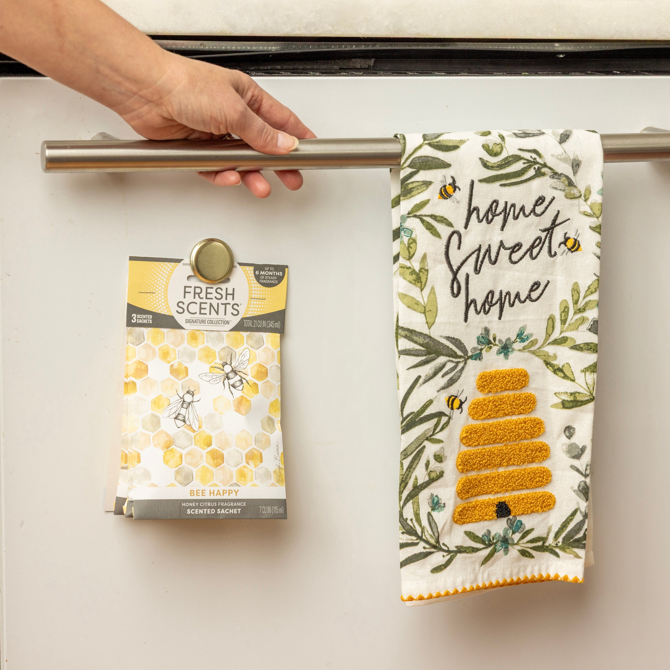 A hand hangs a "Home Sweet Home" bee and floral towel next to Fresh Scents' Bee Happy Sachet 3pk—a flameless air freshener for long-lasting fragrance.