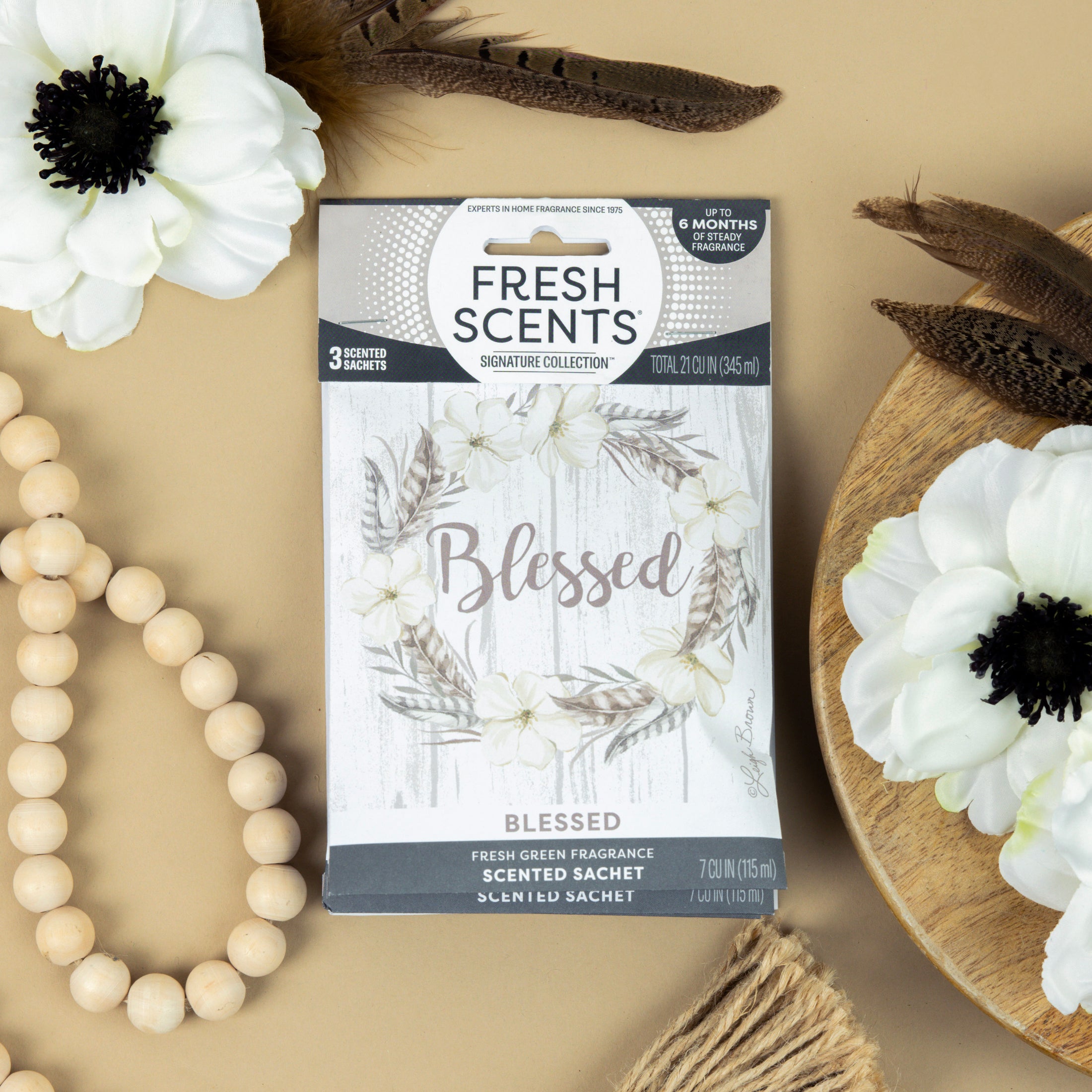 The Fresh Scents Blessed Wreath 3pk scented sachets feature a floral wreath design and the word "Blessed" on the box, shown with white flowers and wooden beads—an ideal flameless fragrance for any space.
