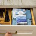 Load image into Gallery viewer, An open kitchen drawer holds folded blue and white dish towels, utensils in a wooden organizer, and a Fresh Scents Adventure Mountain Sachet 3pk for long-lasting freshness. A hand pulls the drawer open beneath a marble countertop.