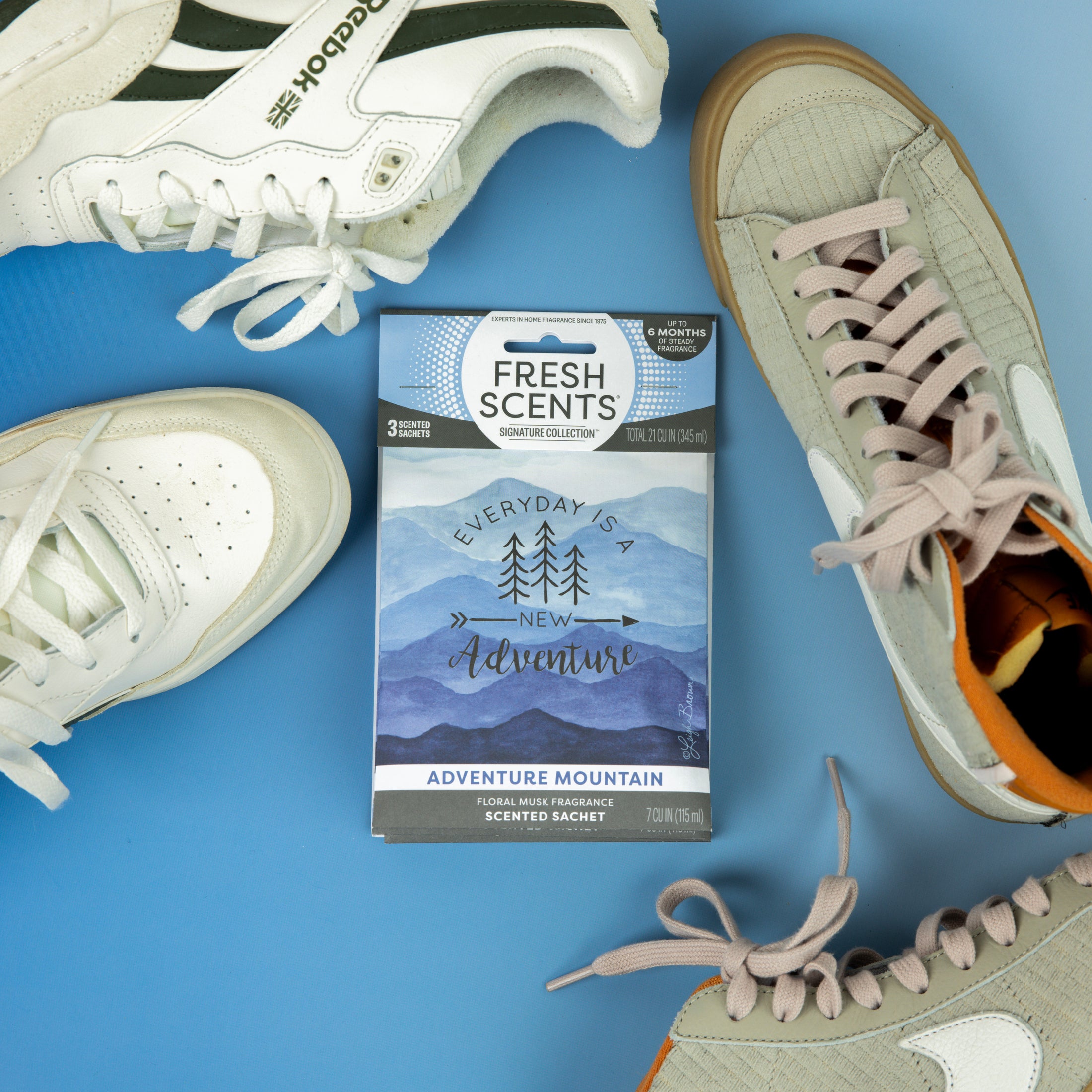 A blue Fresh Scents Adventure Mountain Sachet 3pk lies on a blue surface, surrounded by three pairs of white and beige sneakers, providing long-lasting freshness.