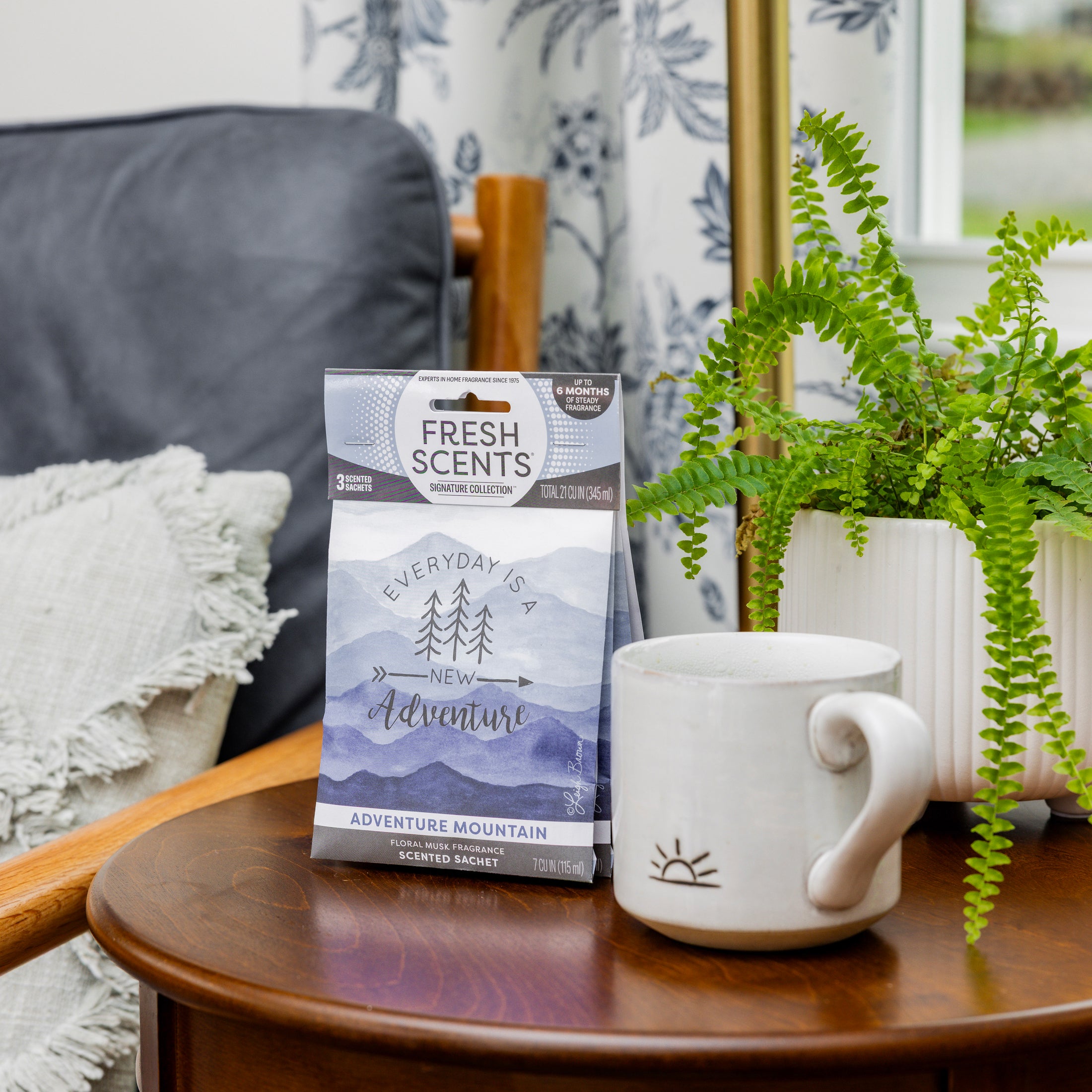 A small table holds a white mug, a potted fern, and a blue bag of Fresh Scents Adventure Mountain - Sachet 3pk for long-lasting freshness. In the background are a gray pillow, wooden chair, and floral curtain by the window.