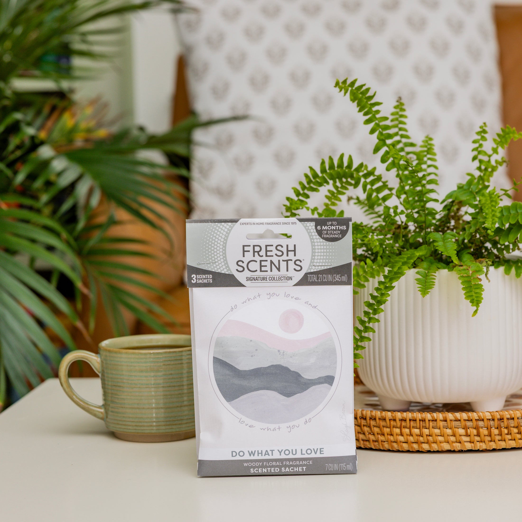 A Fresh Scents "Do What You Love" Sachet 3pk, a flameless fragrance for any space, sits on a white table by a green mug and potted fern. Behind it are a wicker tray, leafy plant, and patterned pillow.