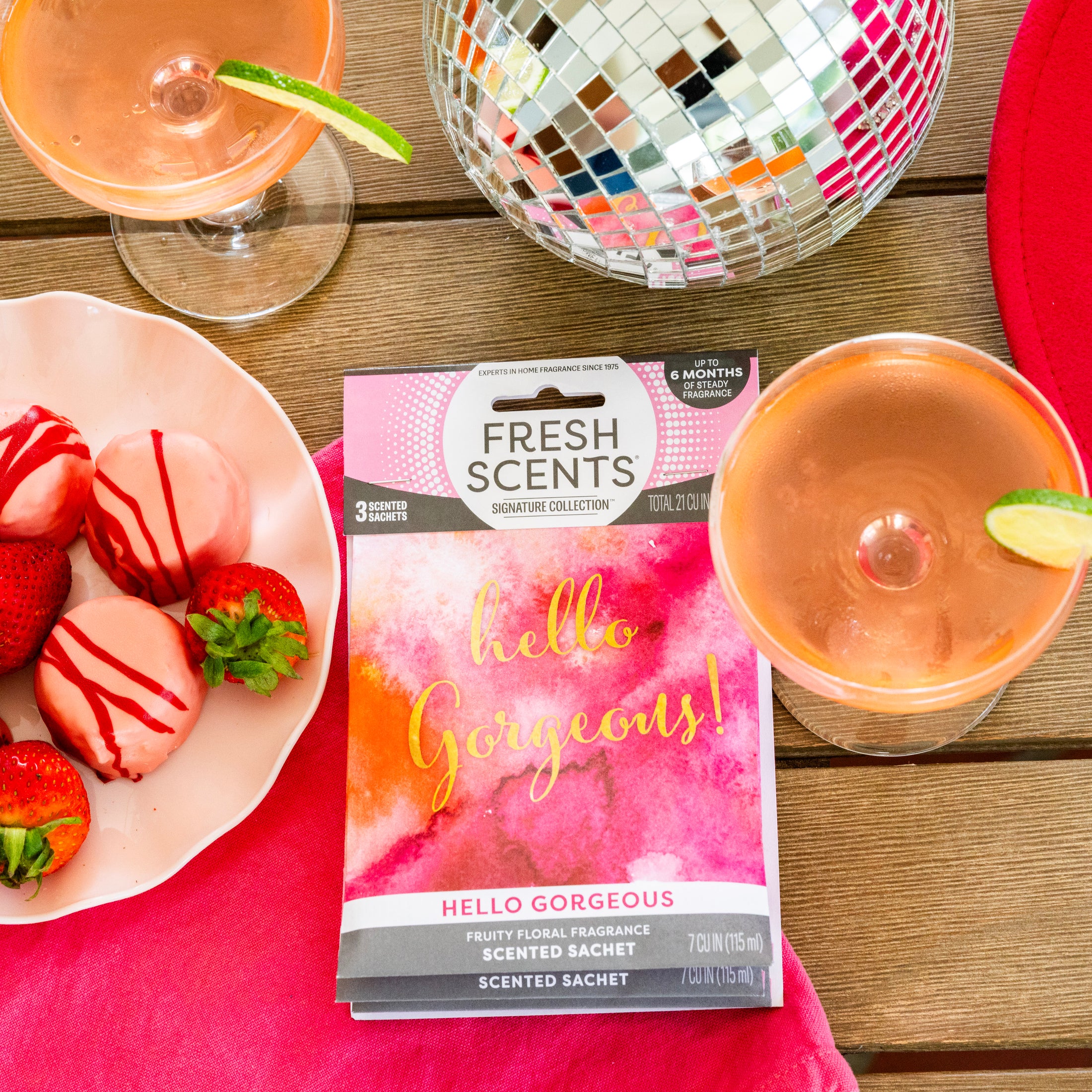 A vibrant flat lay with two pink cocktails, chocolate-covered strawberries, and a Fresh Scents Hello Gorgeous - Sachet 3pk for lasting fragrance, alongside a disco ball on a colorful table setting.