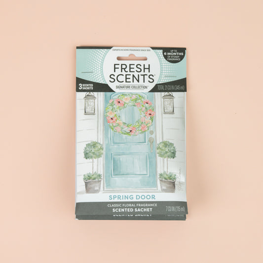 The Fresh Scents Spring Door Sachet 3pk features a flameless fragrance, with packets illustrated with a blue door, floral wreath, and potted plants on a peach background.