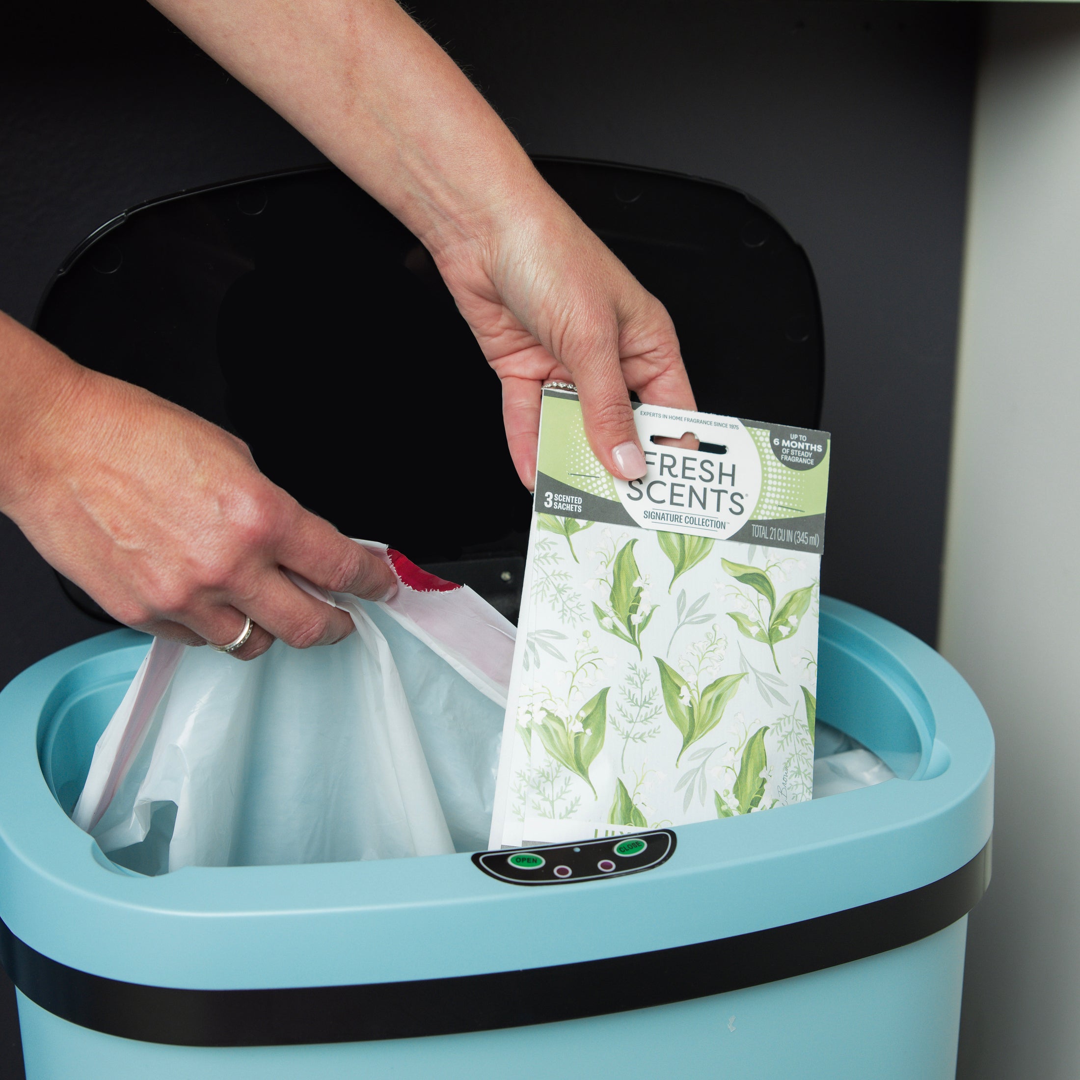 A person places a Fresh Scents Lily of the Valley Sachet 3pk, a flameless air freshener, and a white trash bag into a blue trash bin with a black lid.