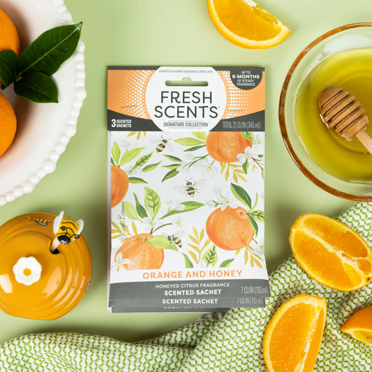 A Fresh Scents Orange & Honey - Sachet 3pk, a long-lasting freshener, is surrounded by orange slices, a honey dipper in honey, a yellow teapot, green leaves, and a white dish on a light green surface.