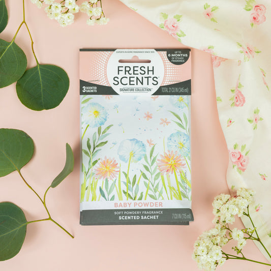 A package of Fresh Scents Baby Powder - Sachet 3pk, a long-lasting air freshener, lies on a pink surface with green leaves and baby's breath flowers. The packaging features pastel flower illustrations and floral-patterned cloth accents.