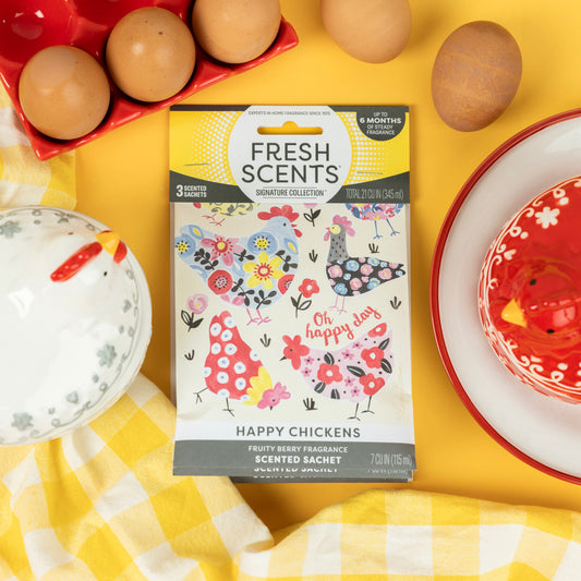 The Fresh Scents Happy Chicken - Sachet 3pk, featuring flower and chicken designs, rests on a yellow surface with brown eggs, a red dish, a chicken-shaped ceramic dish, and a yellow checkered cloth for lasting fragrance.