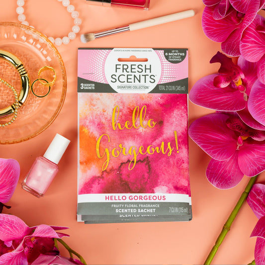 A Fresh Scents Hello Gorgeous - Sachet 3pk flameless air freshener with a bright pink and orange watercolor design rests on a pink surface among orchids, pearls, gold jewelry, and nail polish, promising long-lasting fragrance.