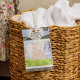 Load image into Gallery viewer, A woven laundry basket filled with white towels has a Fresh Scents Clean Clothes - Sachet 3pk clipped to the front for flameless fragrance. A floral dress hangs nearby.
