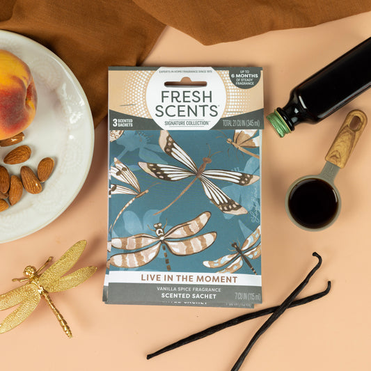 A box of Fresh Scents Live in the Moment - Sachet 3pk rests on a tan surface, surrounded by a peach, almonds, vanilla beans, a gold dragonfly, black bottle, and measuring cup for long-lasting flameless fragrance.