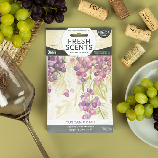 A Fresh Scents Tuscan Grape - Sachet 3pk, a flameless air freshener with long-lasting fragrance, sits on a green surface beside red wine, corks, and plates of green and purple grapes.