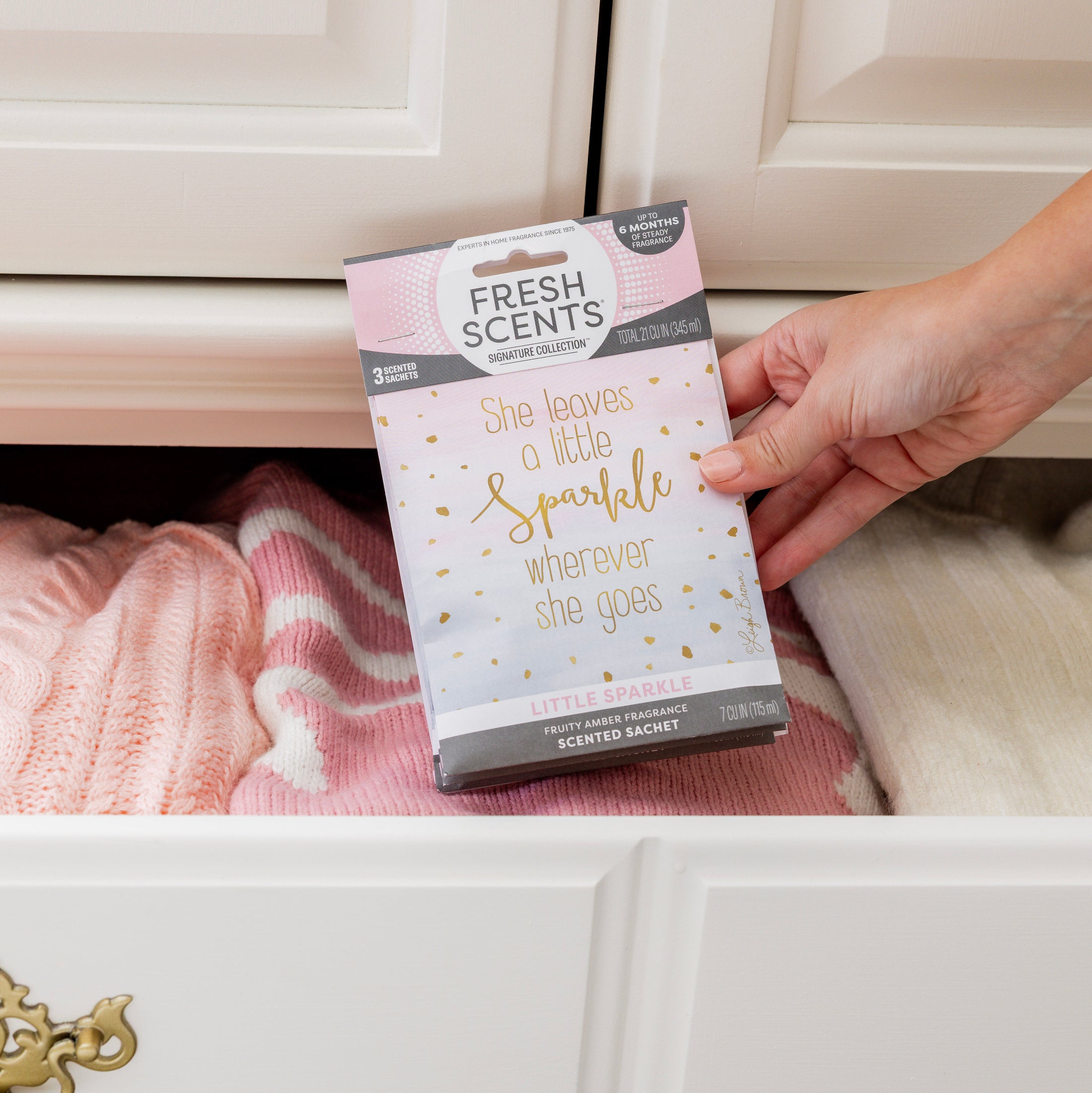 A hand places a Fresh Scents Little Sparkle - Sachet 3pk with the quote "She leaves a little sparkle wherever she goes" into a drawer of folded pink and cream sweaters, giving your wardrobe a long-lasting scent.