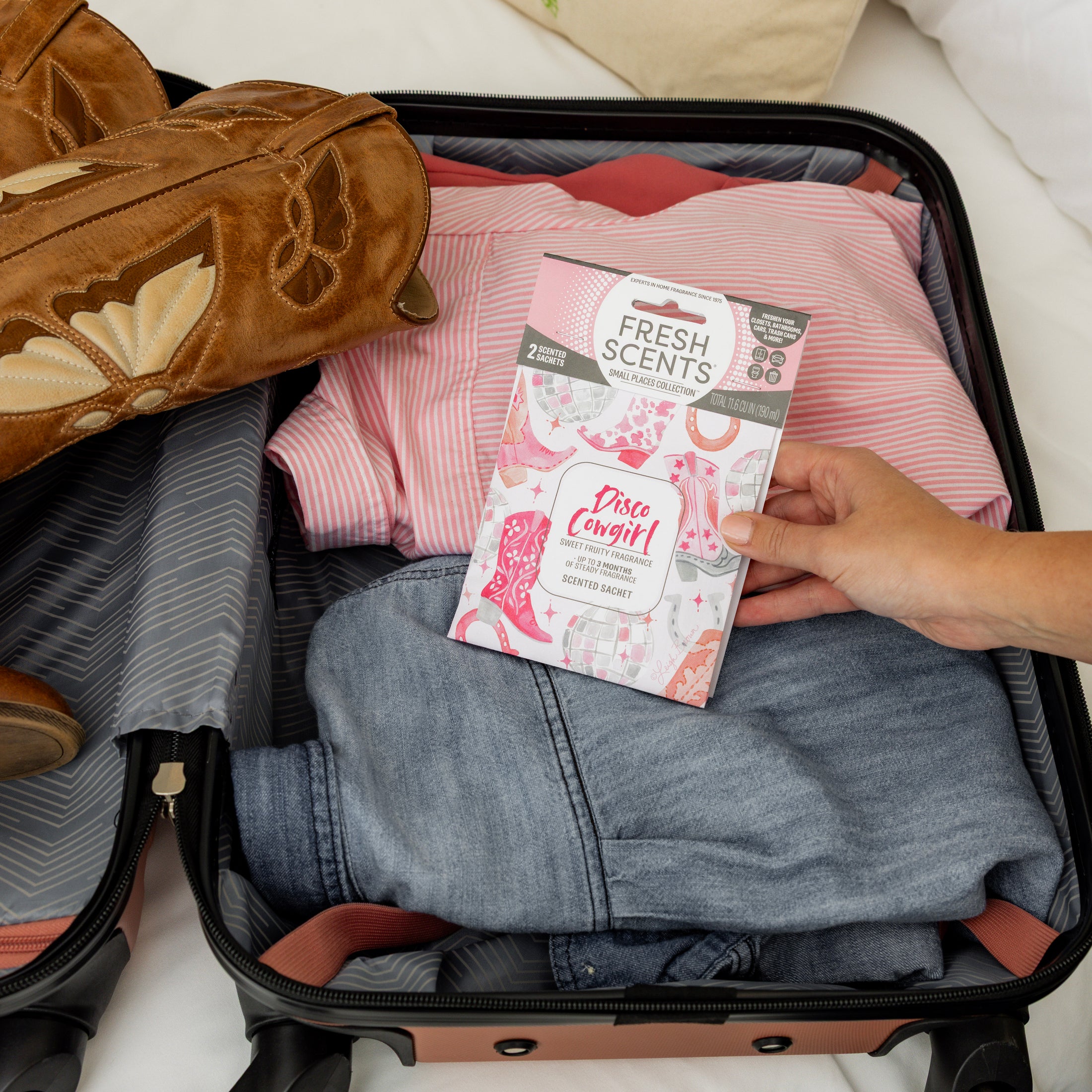 A hand packs the Fresh Scents Disco Cowgirl - Sachet 2pk into a suitcase with shirts, jeans, and cowboy boots, adding travel-ready fragrance for small spaces.