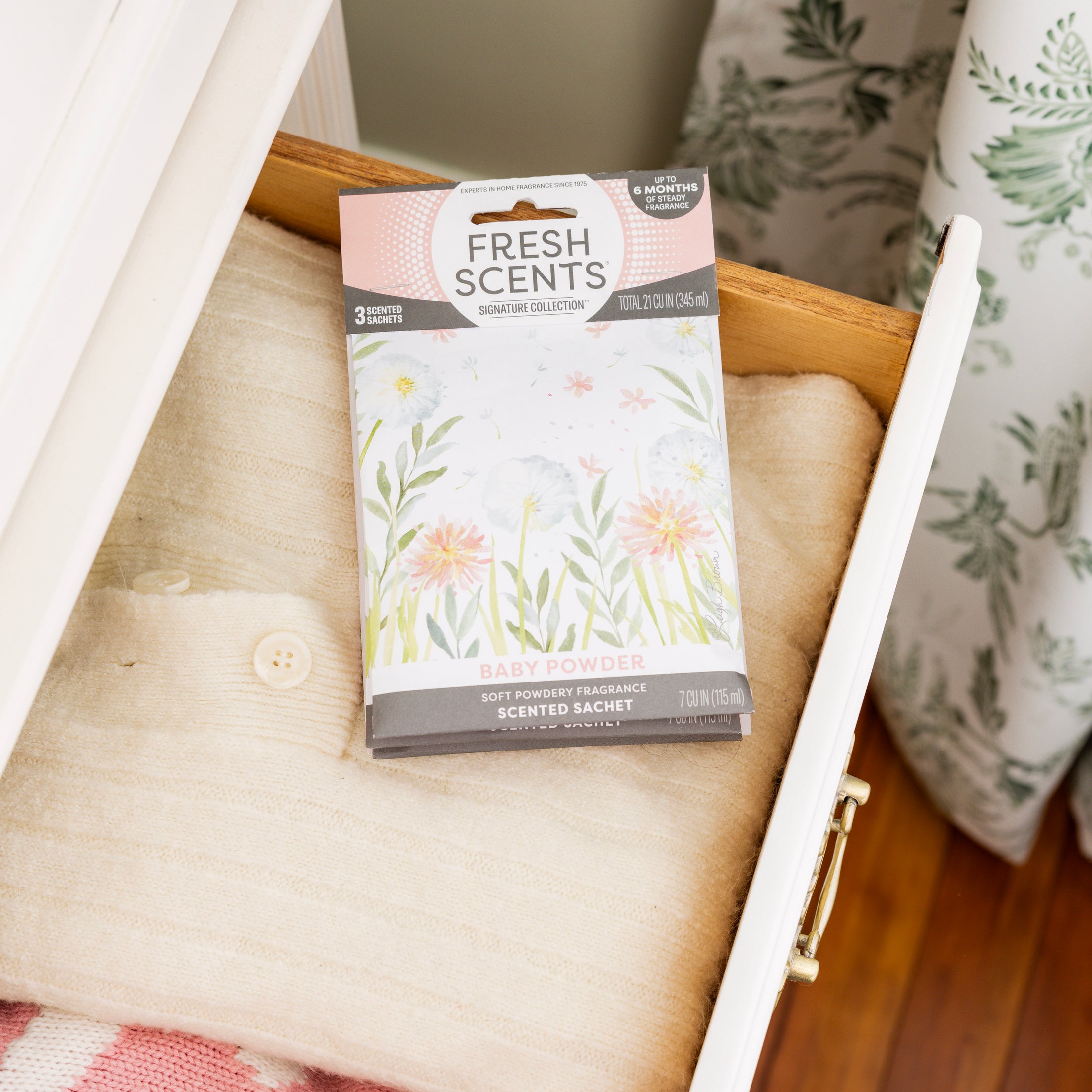 The Fresh Scents Baby Powder - Sachet 3pk offers a flameless fragrance option, shown resting on sweaters in a partly open white drawer with floral curtains in the background.