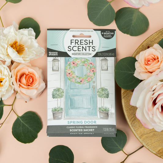 A Fresh Scents Spring Door - Sachet 3pk, a long-lasting air freshener, sits on a peach surface with pink roses and eucalyptus nearby. The packaging features an illustrated blue door with a floral wreath. One bouquet rests on a wooden tray.