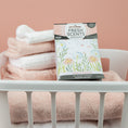 Load image into Gallery viewer, A white laundry basket with folded pink and white towels holds a Fresh Scents Baby Powder - Sachet 3pk, providing long-lasting fragrance against a pale pink background.
