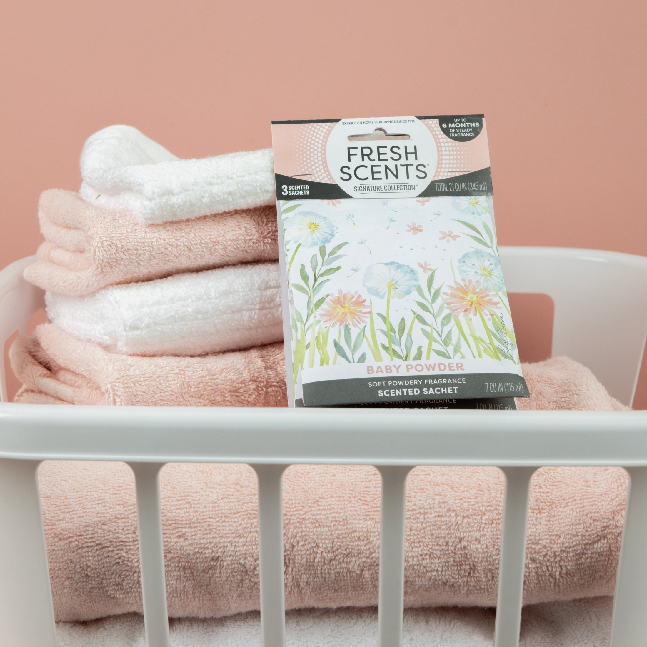 A white laundry basket with folded pink and white towels holds a Fresh Scents Baby Powder - Sachet 3pk, providing long-lasting fragrance against a pale pink background.
