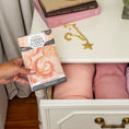 Load image into Gallery viewer, A hand puts a Fresh Scents Summer Romance - Sachet 3pk, a flameless, long-lasting air freshener, into a drawer of pink and peach fabrics. Books and a gold star necklace rest on top of the dresser.
