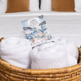 Load image into Gallery viewer, A woven basket with rolled white towels and a Fresh Scents Painted Palm - Sachet 3pk, a flameless fragrance option, rests on a white bed with two brown pillows in the background.
