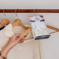 Load image into Gallery viewer, A hand hangs a cream knit sweater and a Fresh Scents Baby Powder - Sachet 3pk, a flameless fragrance, from a wooden hanger on a closet rod.
