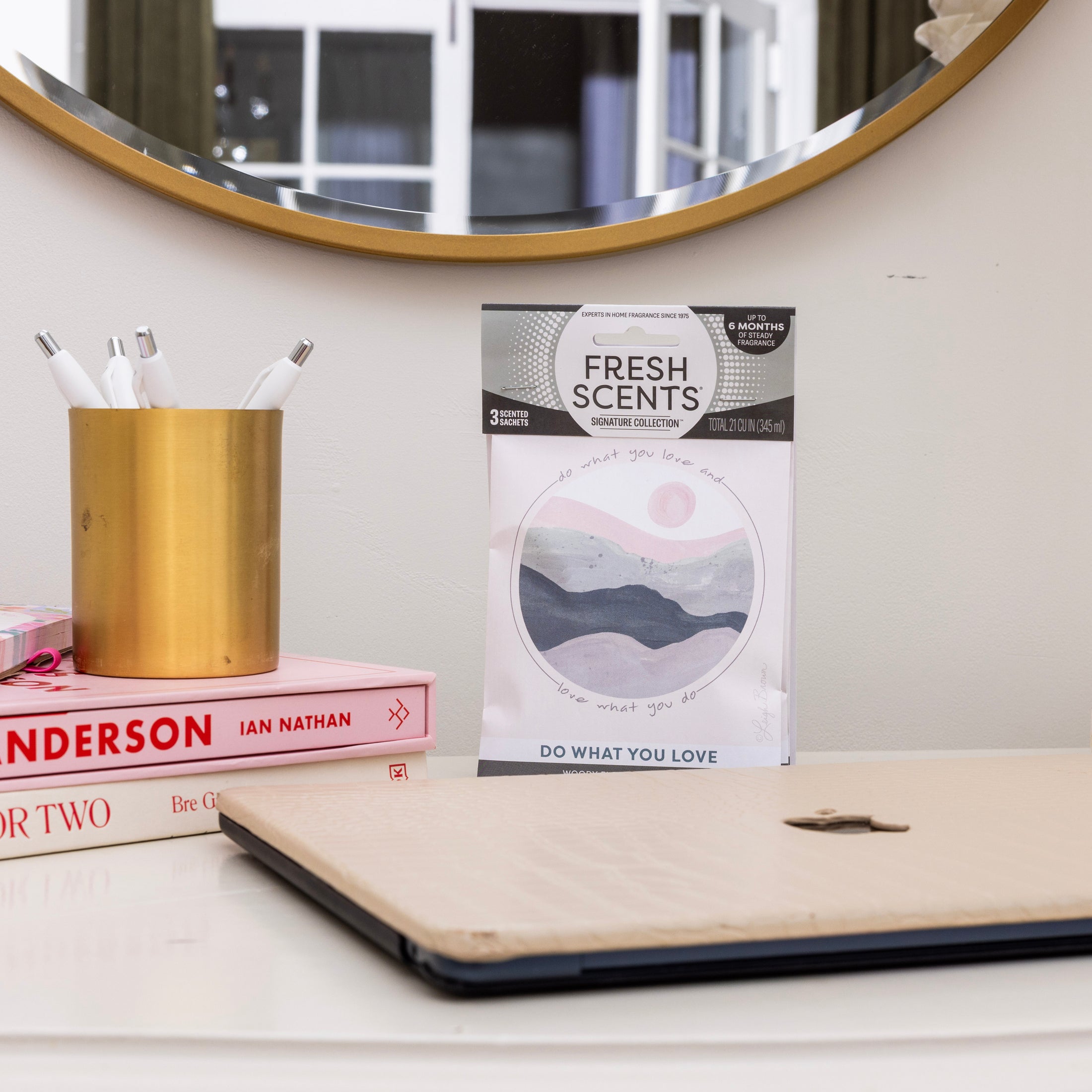 A desk with a closed laptop, books, a gold pen holder, and a Fresh Scents Do What You Love - Sachet 3pk in front of a round mirror—a stylish, flameless way to keep your room fresh for longer.