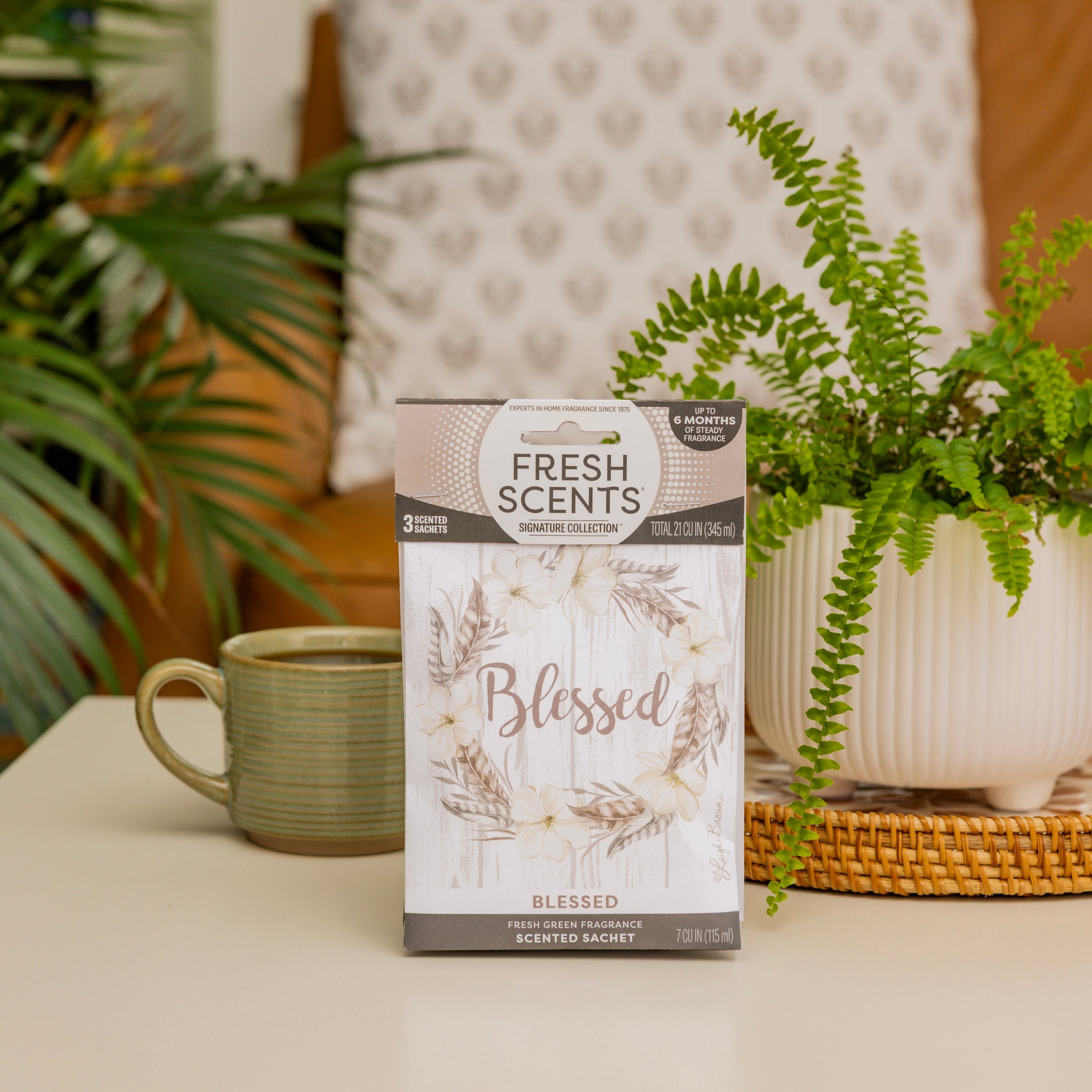 A box labeled Fresh Scents Blessed Wreath 3pk, a long-lasting air freshener, sits on a table with a green fern in a white pot, a light green mug, and wicker decor, while leafy plants and a patterned pillow are in the background.