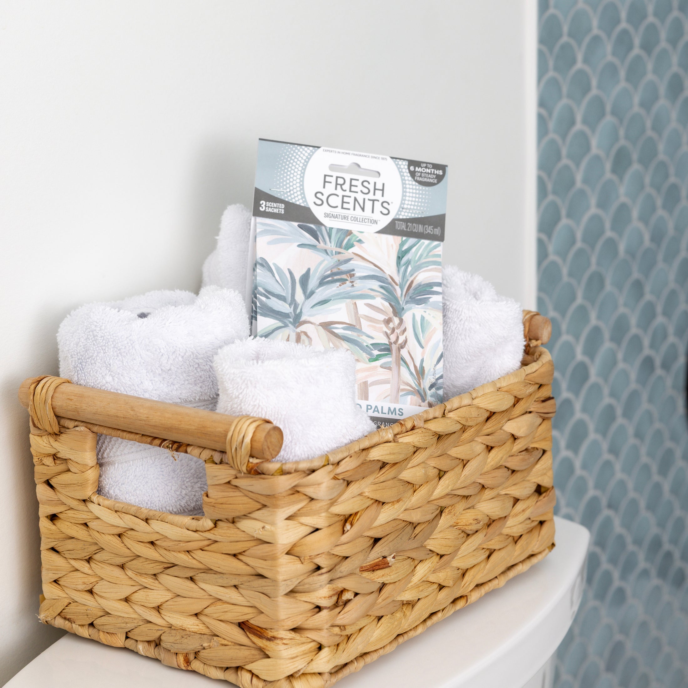 A woven basket with rolled white towels and Fresh Scents Painted Palm - Sachet 3pk sits on a white surface before blue patterned tiles, adding a flameless fragrance to the space.