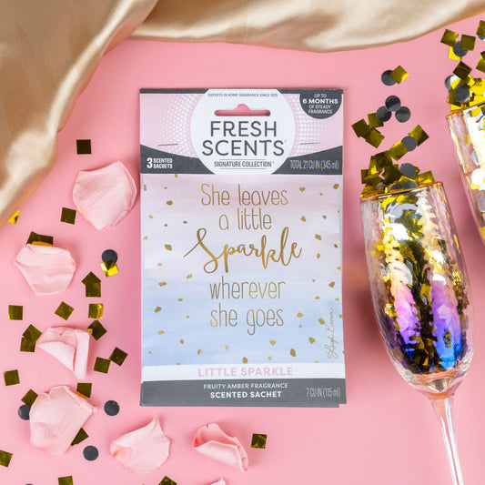 The Fresh Scents Little Sparkle - Sachet 3pk, featuring “She leaves a little sparkle wherever she goes,” rests on pink with rose petals, gold confetti, and a champagne flute—a flameless air freshener with long-lasting fragrance.