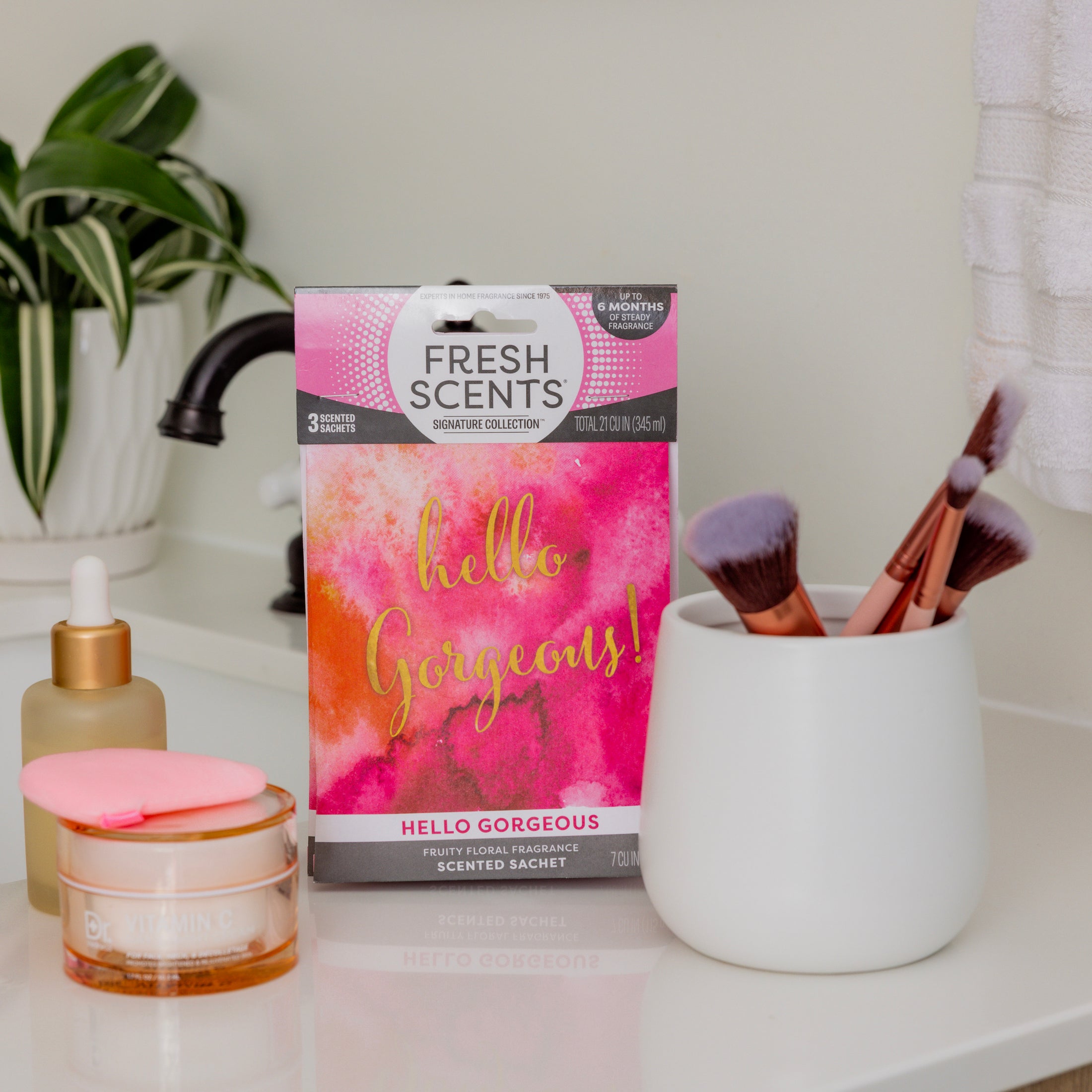 A pink and white Hello Gorgeous - Sachet 3pk by Fresh Scents sits on a bathroom counter, providing long-lasting fragrance beside a makeup brush cup, glass dropper bottle, and a cream jar with a pink applicator.