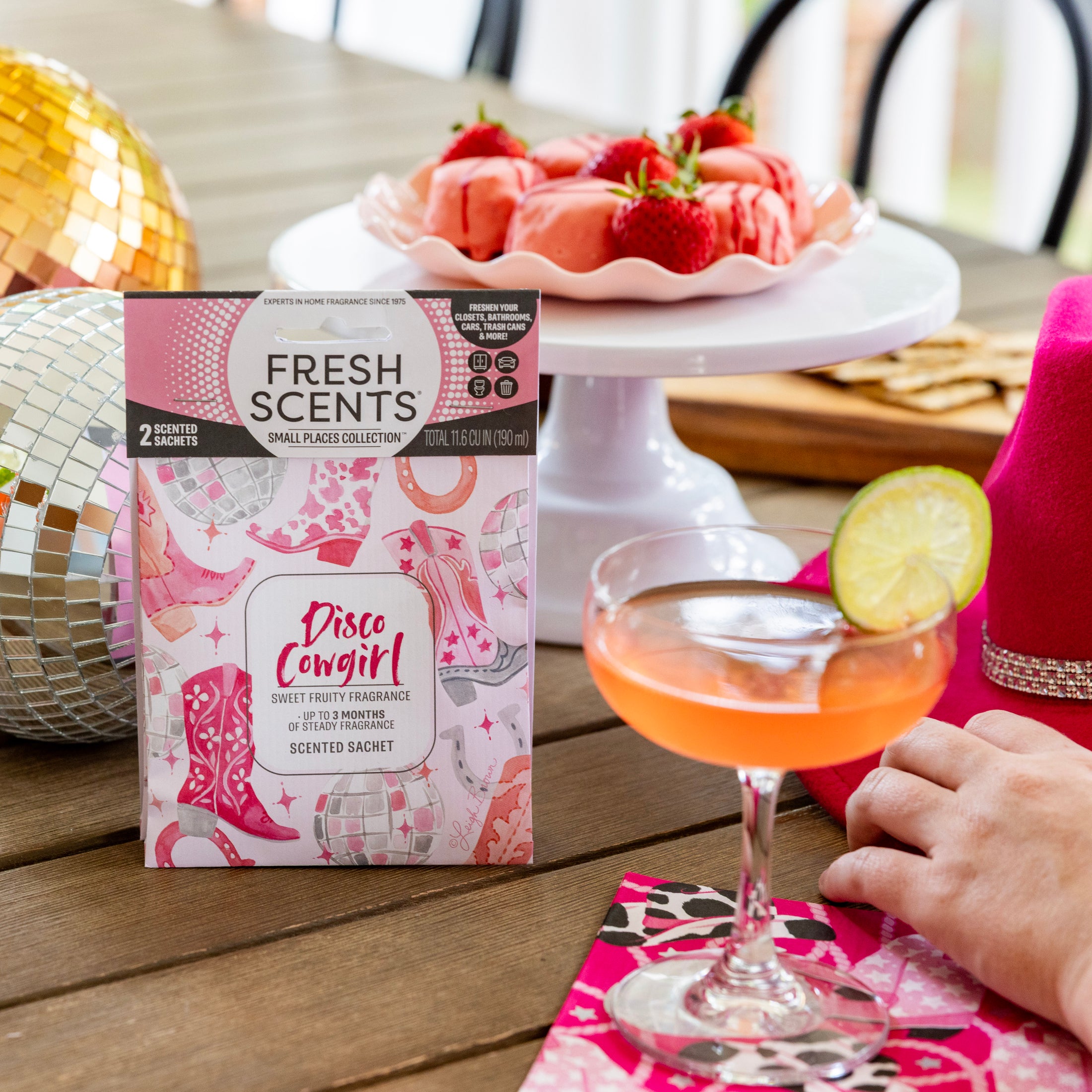 A pink cocktail with a lime slice sits on a wooden table next to a hand, pink napkin, Fresh Scents Disco Cowgirl Sachet 2pk box, chocolate-covered strawberries, a pink hat, and a sparkling disco ball.