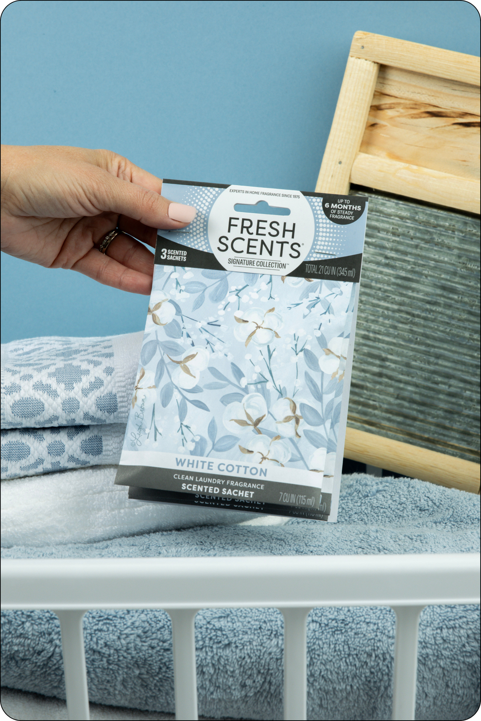A hand holds a Fresh Scents White Cotton scented sachet above folded towels and a laundry washboard in a basket, set against a light blue background.